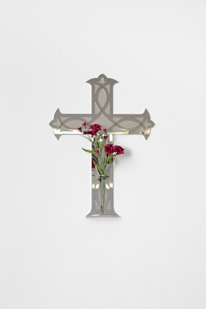Wandskulptur: spiegelndes Metallkreuz mit Glasvase und roten Nelken an weißer Wand.