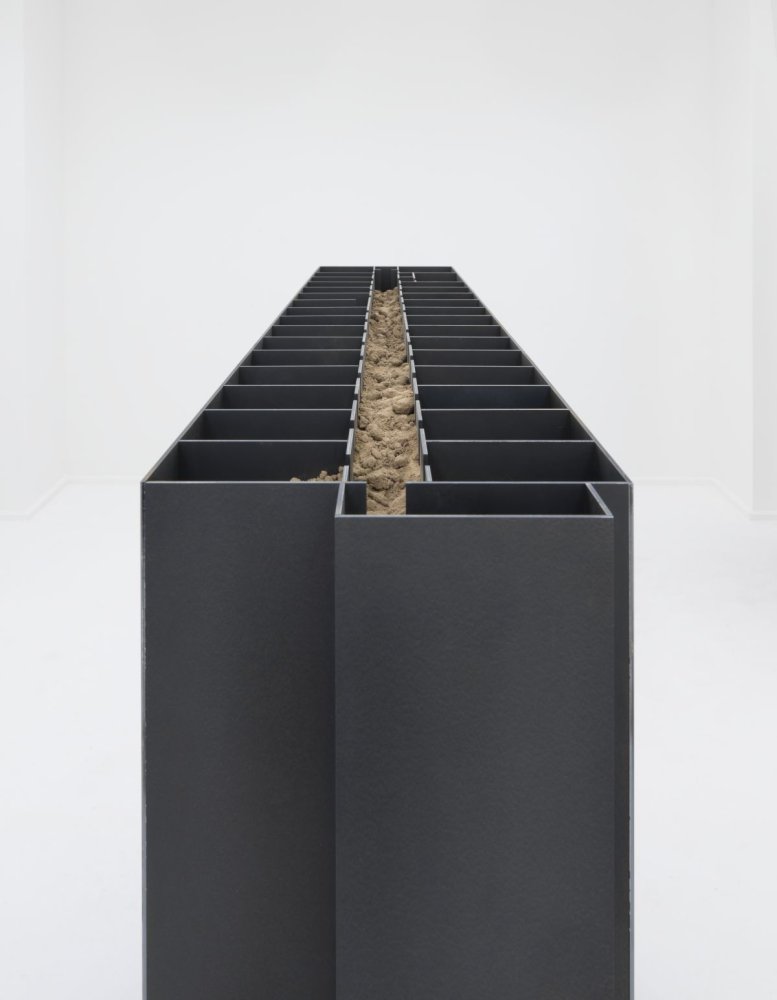 Installationsansicht: Reihe schwarzer Metallkanäle; zentrales Fach mit Erde, im weißen Galerieraum