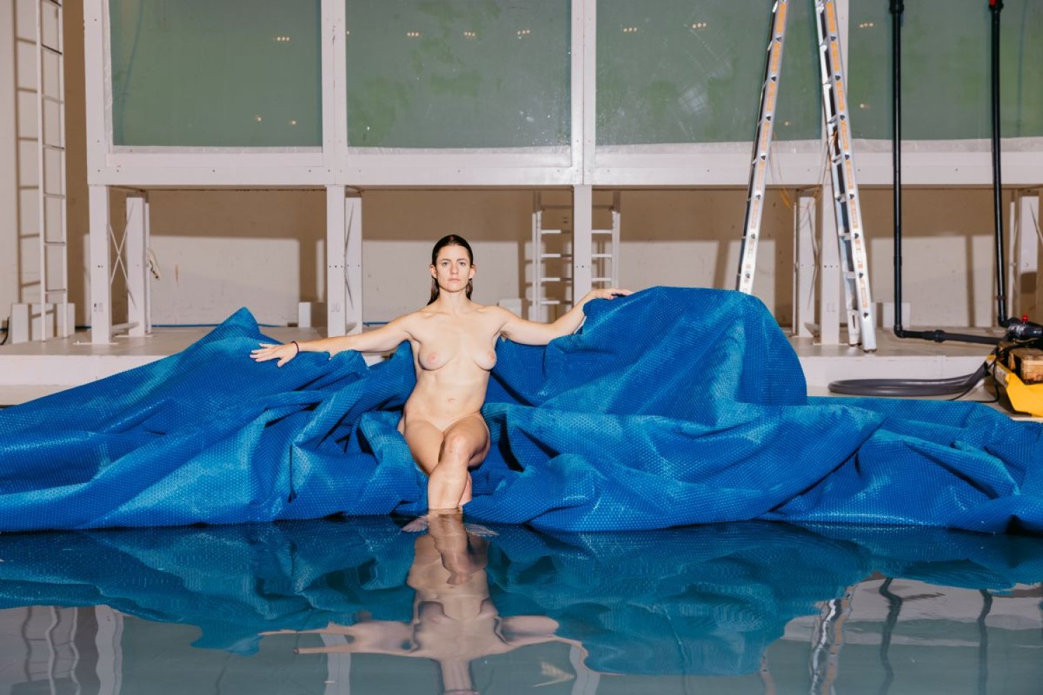 Fotografie: Nackte Person auf blauer Plane am Hallenbadbecken, Leiter im Hintergrund, Spiegelung.