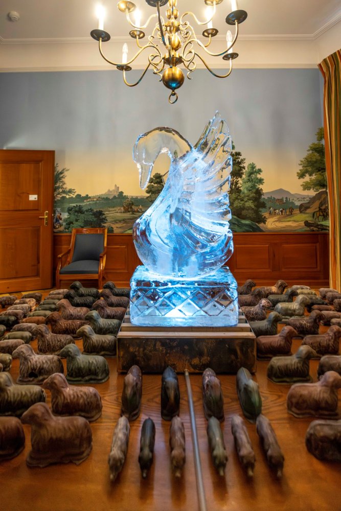 Installationsansicht: Eisschwan auf Holztisch, umgeben von kleinen Objekten; Salon mit Wandbild.