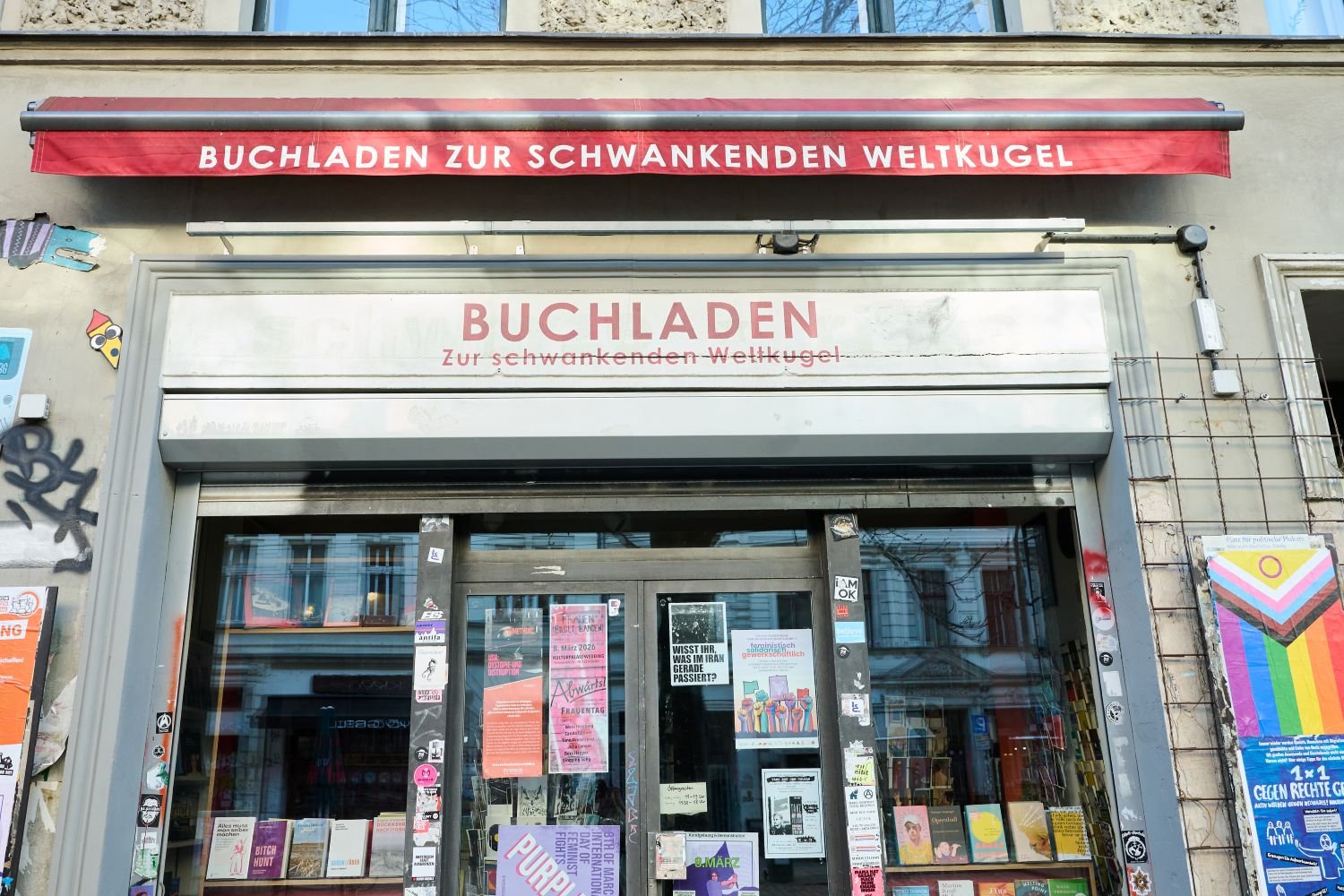 Foto, Fassade des 'Buchladen zur Schwankenden Weltkugel', gläserner, plakatierter Eingang.