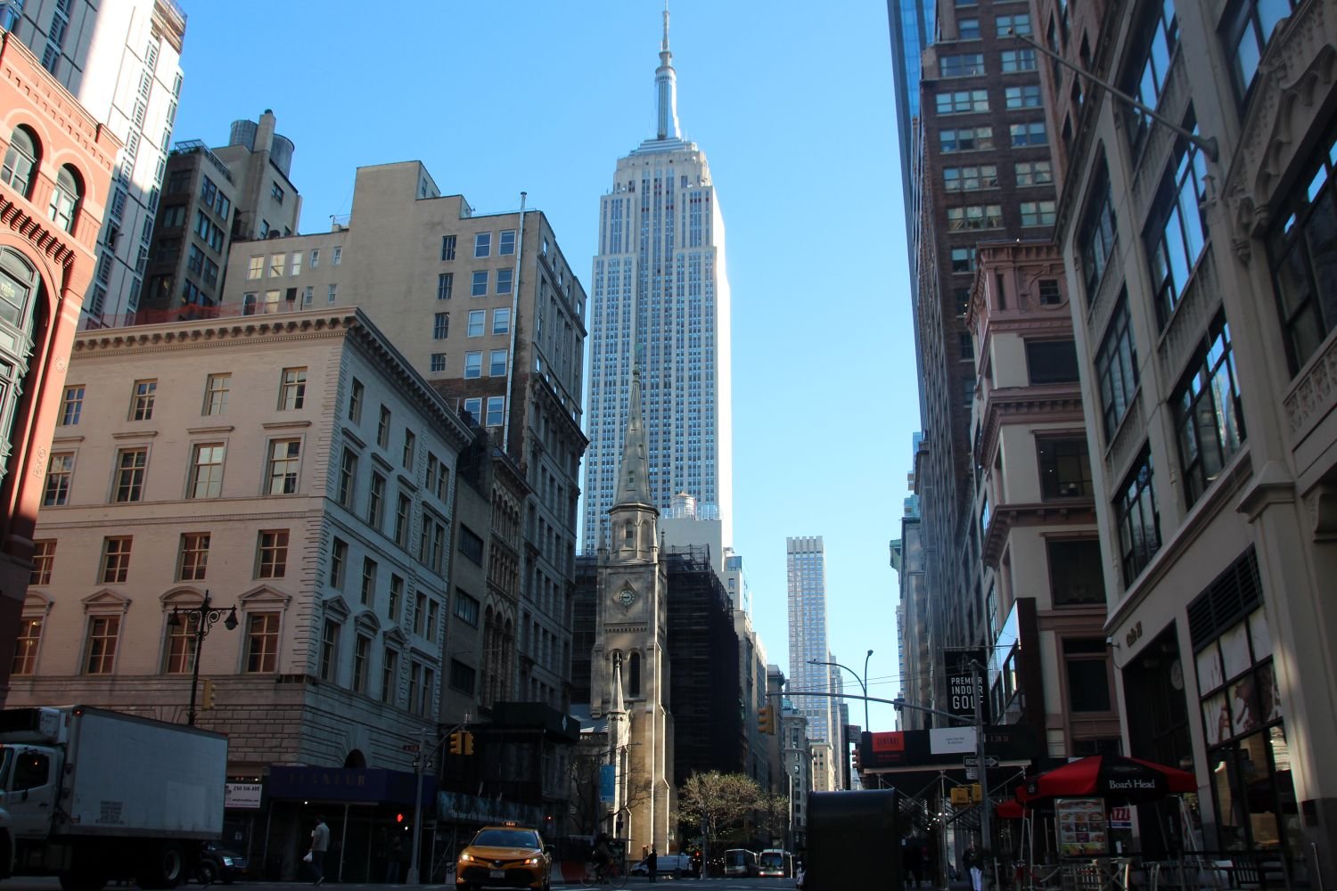 Foto: Empire State Building aus Straßenperspektive in Manhattan, zwischen Altbauten, klarer Himmel.