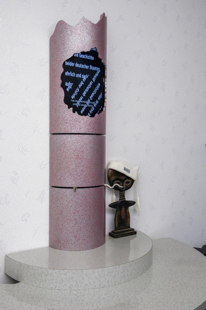 Installationsansicht: pinke Säule mit Ausriss/Text; kleine Büste mit Stoff
