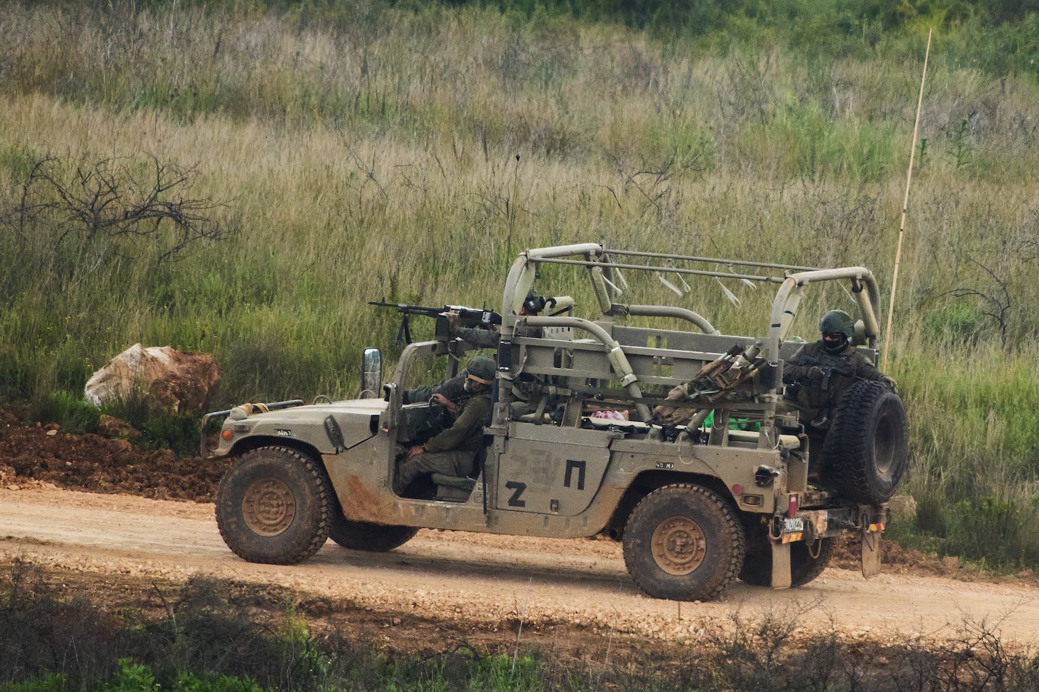 Foto: Militärgeländewagen mit zwei Soldaten auf Erdstraße in grasiger Hügellandschaft.