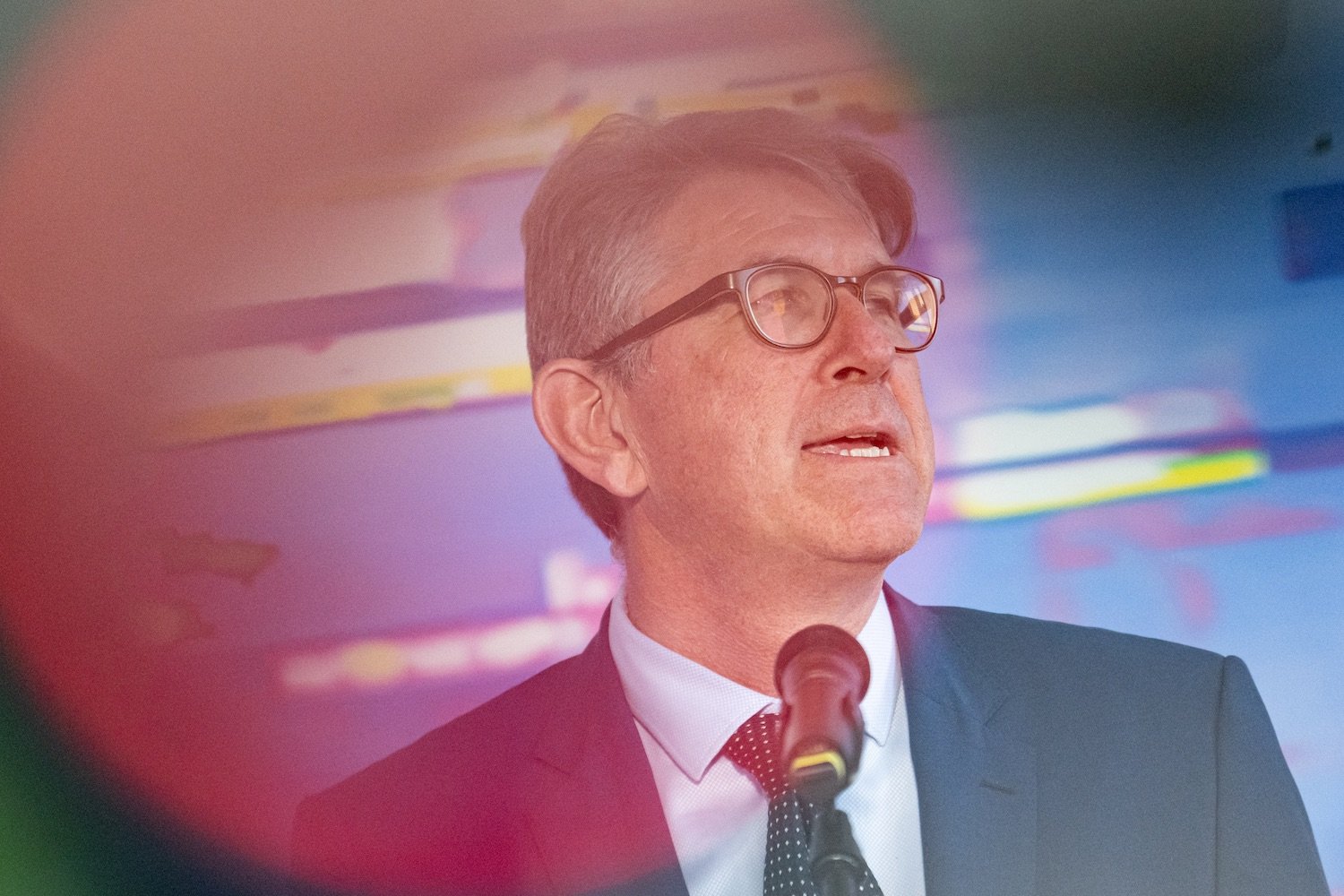 Redner im Anzug am Mikrofon auf Bühne, bunter unscharfer Hintergrund.