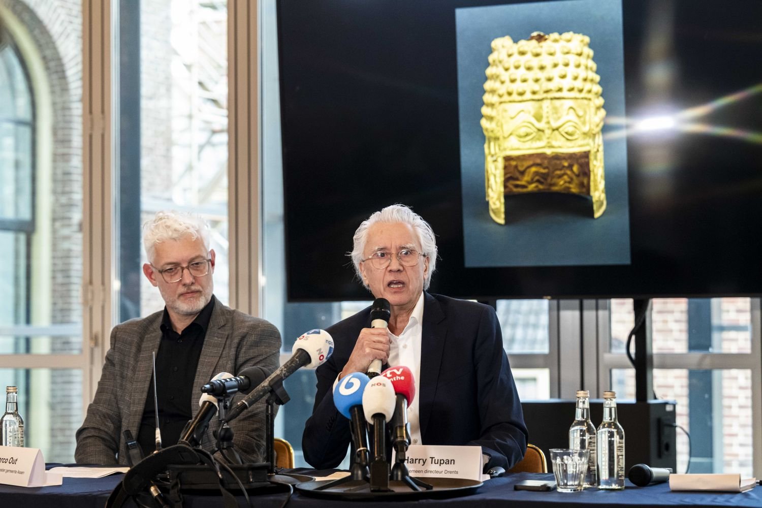 Pressekonferenz im Museum: zwei Sprecher am Tisch; Bildschirm zeigt goldenes Artefakt.