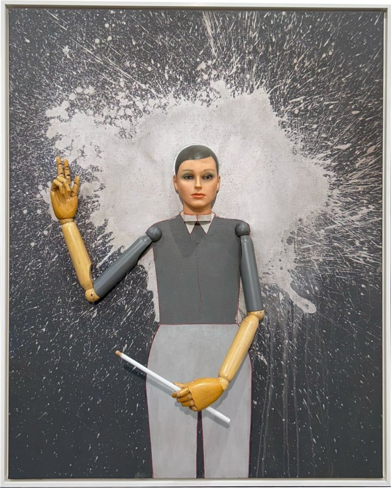 Puppenfigur mit Holzarmen, Stift in rechter Hand, vor dunkler Wand mit weißer Spritzspur, gerahmt.