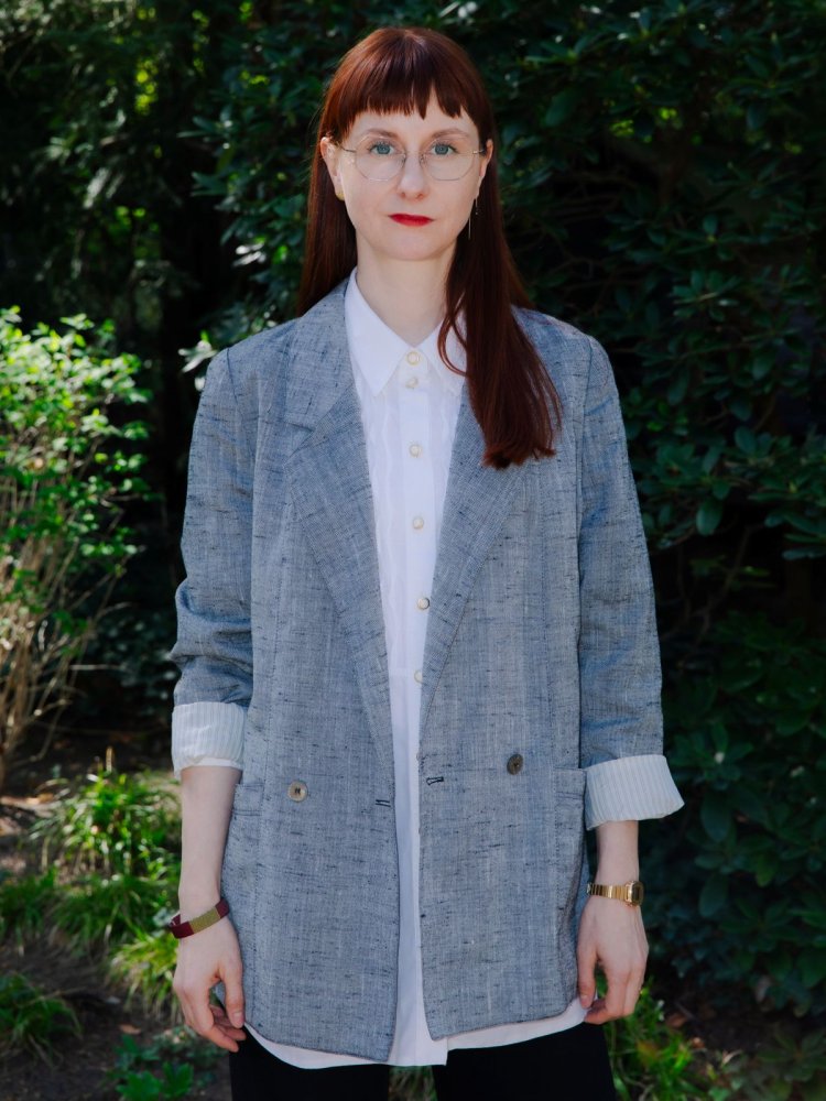 Porträt einer Person mit langem rotem Haar, frontal im Garten; grauer Blazer, neutraler Blick.