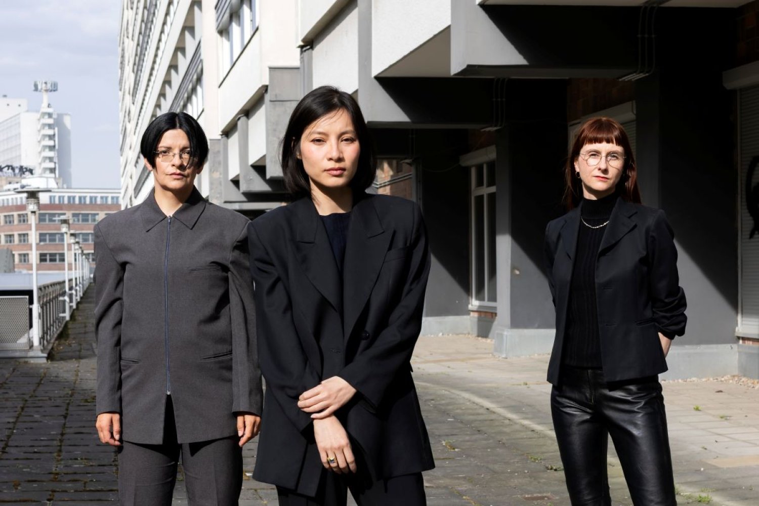 Foto, Gruppenporträt: Drei Personen in dunklen Anzügen vor Betonbau; eine vorn, blicken zur Kamera.