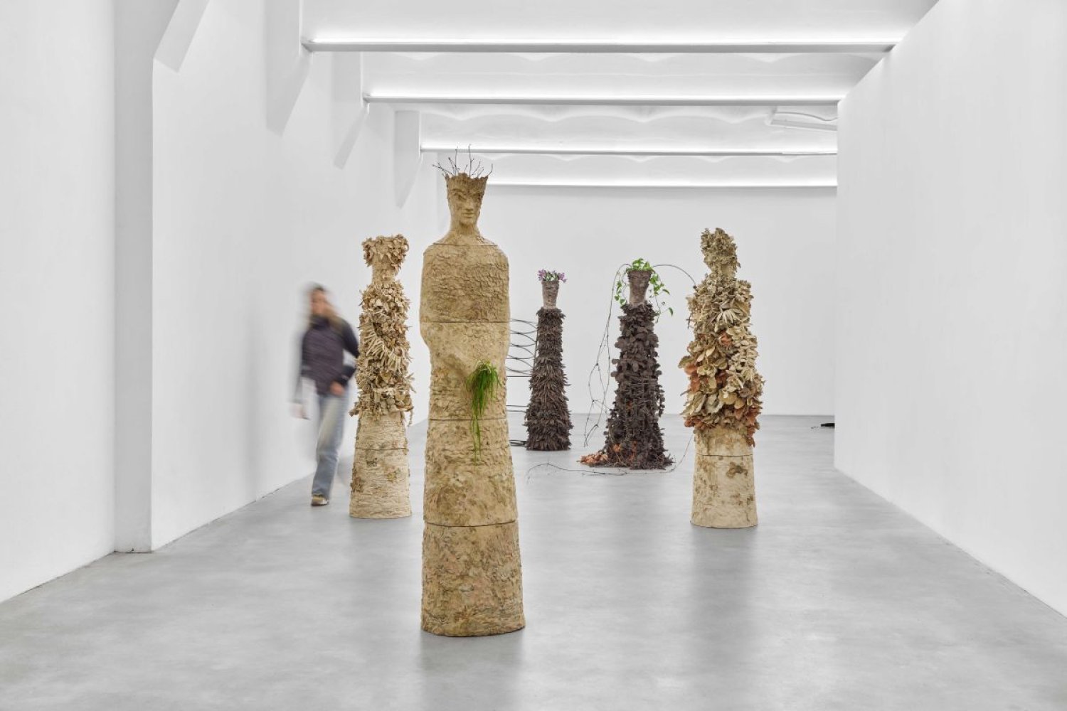 Installationsansicht: menschenhohe Skulpturen aus Naturmaterialien in weißem Raum, versetzt gestellt