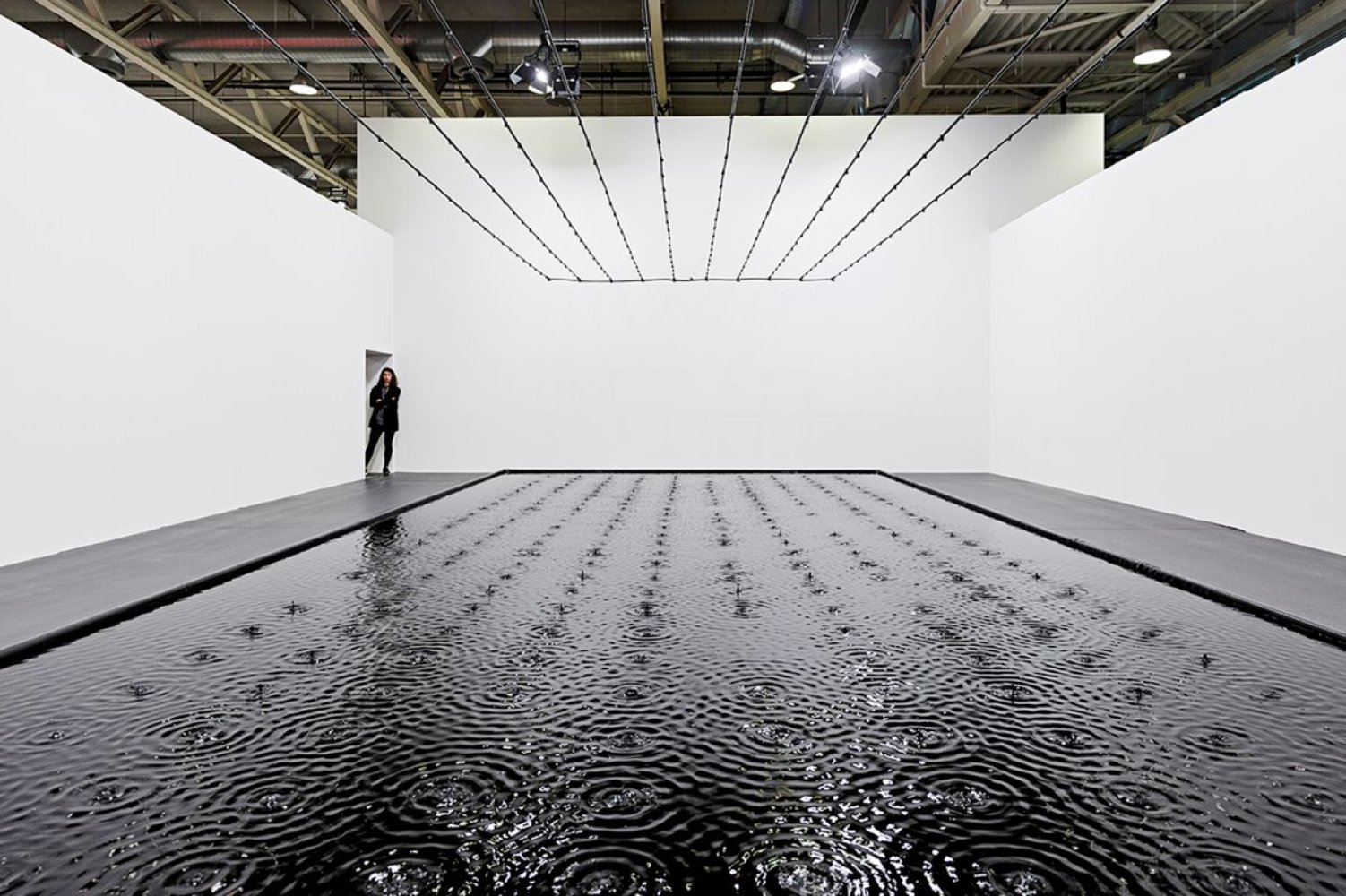 Installationsansicht: großer flacher Wasserpool mit Kreiswellen; weiße Halle, Person hinten links.