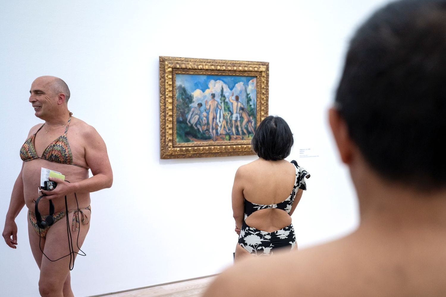 Fotografie: Drei Gäste in Badekleidung betrachten ein goldgerahmtes Gemälde im weißen Galerieraum.