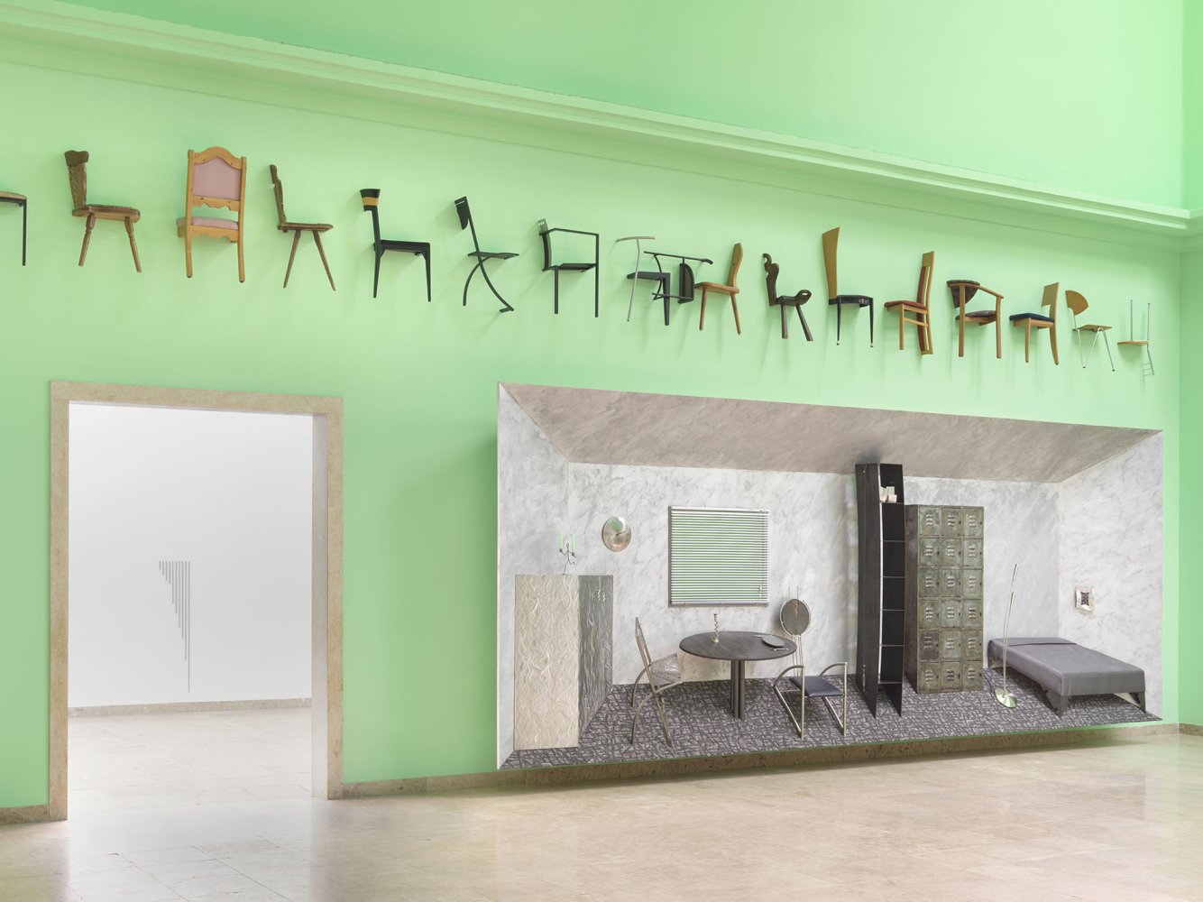 Installationsansicht: grüne Wand, oben Stuhlreihe; großformatige Zimmerkulisse mit Tisch und Bett.