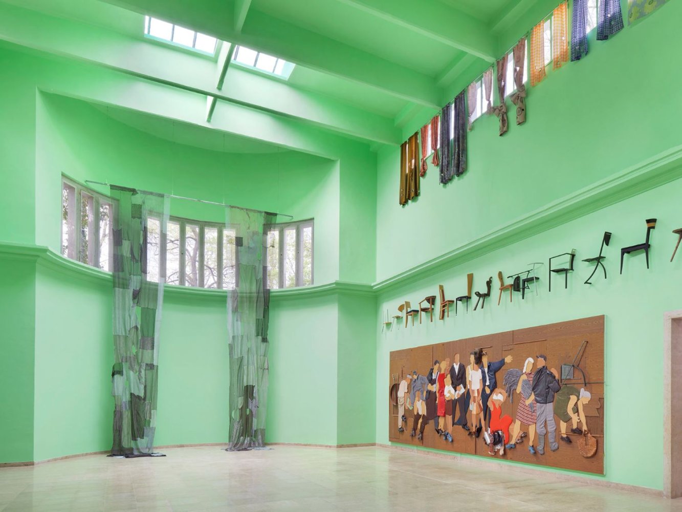 Installationsansicht im hohen grünen Saal: Stühle an der Wand, Wandbild, hängende Paneele