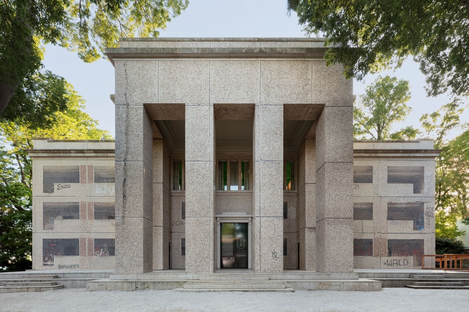 Deutscher Pavillon, Außenansicht; hohe Pfeiler, symmetrische Steinfassade, Bäume im Hintergrund.