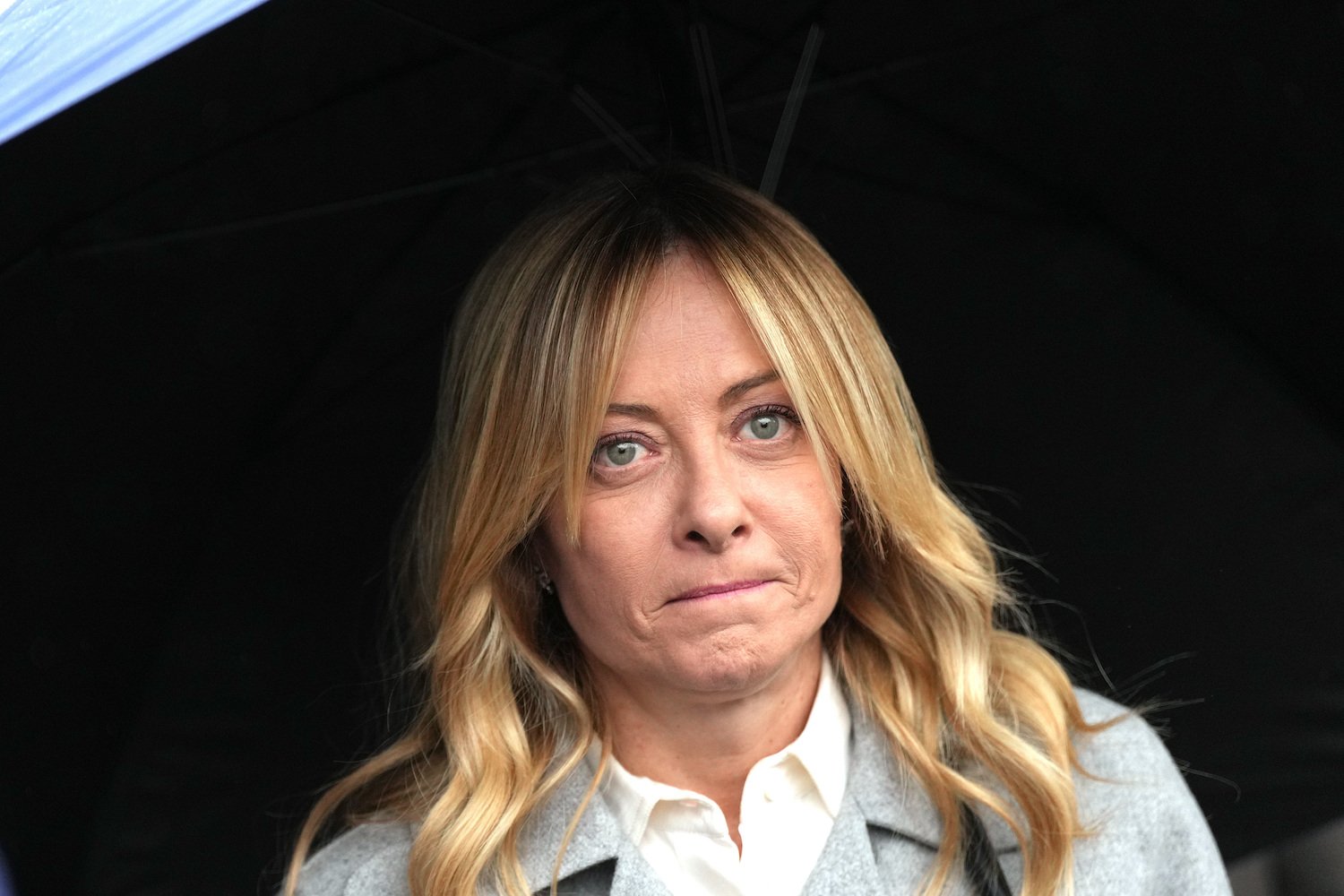 Porträtfoto einer Person unter schwarzem Regenschirm, frontal, blondem Haar, grauer Mantel.