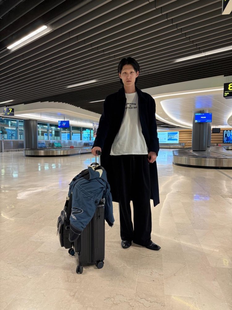 Foto: Person in dunklem Mantel am Flughafen-Gepäckband mit Trolley und Jacke, Blick in die Kamera.