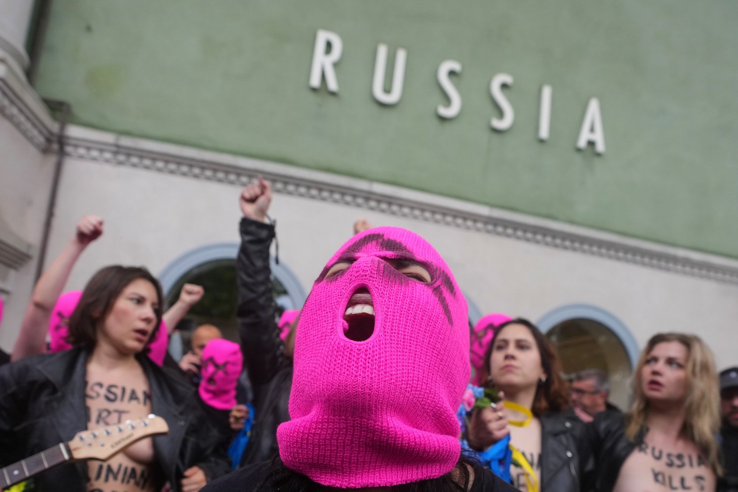 Protestierende in pinken Sturmhauben, Fäuste erhoben, vor Gebäude mit Schriftzug RUSSIA.