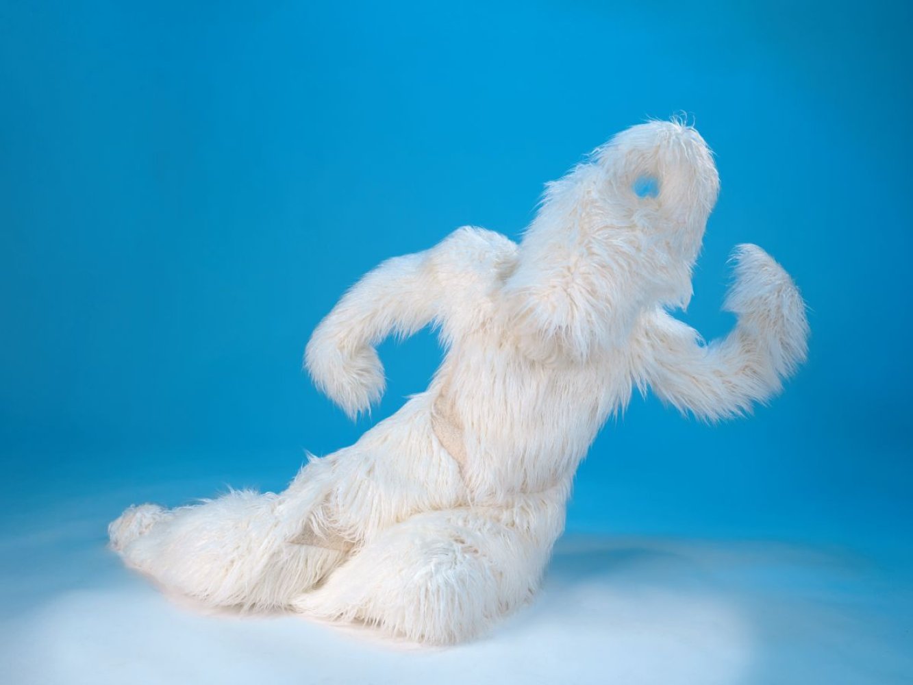 Studioaufnahme einer weißen, fellbedeckten Figur in gebeugter Pose vor hellblauem Studiohintergrund.