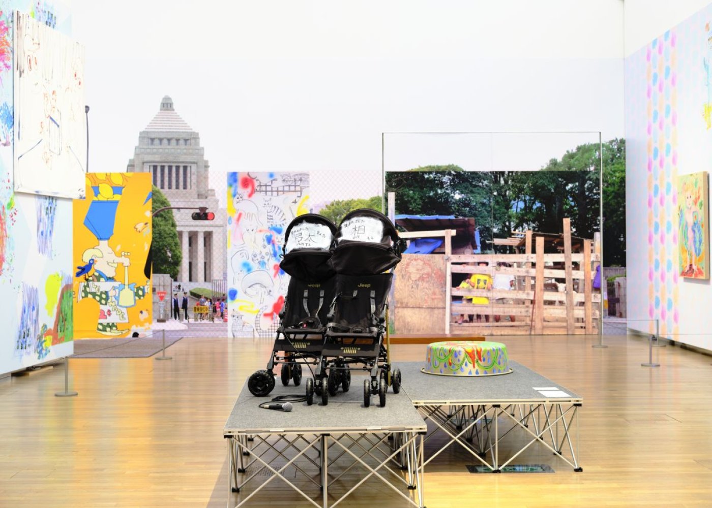 Installationsansicht: zwei Kinderwagen auf Podest, umgeben von großen Fotos und bunten Leinwänden.