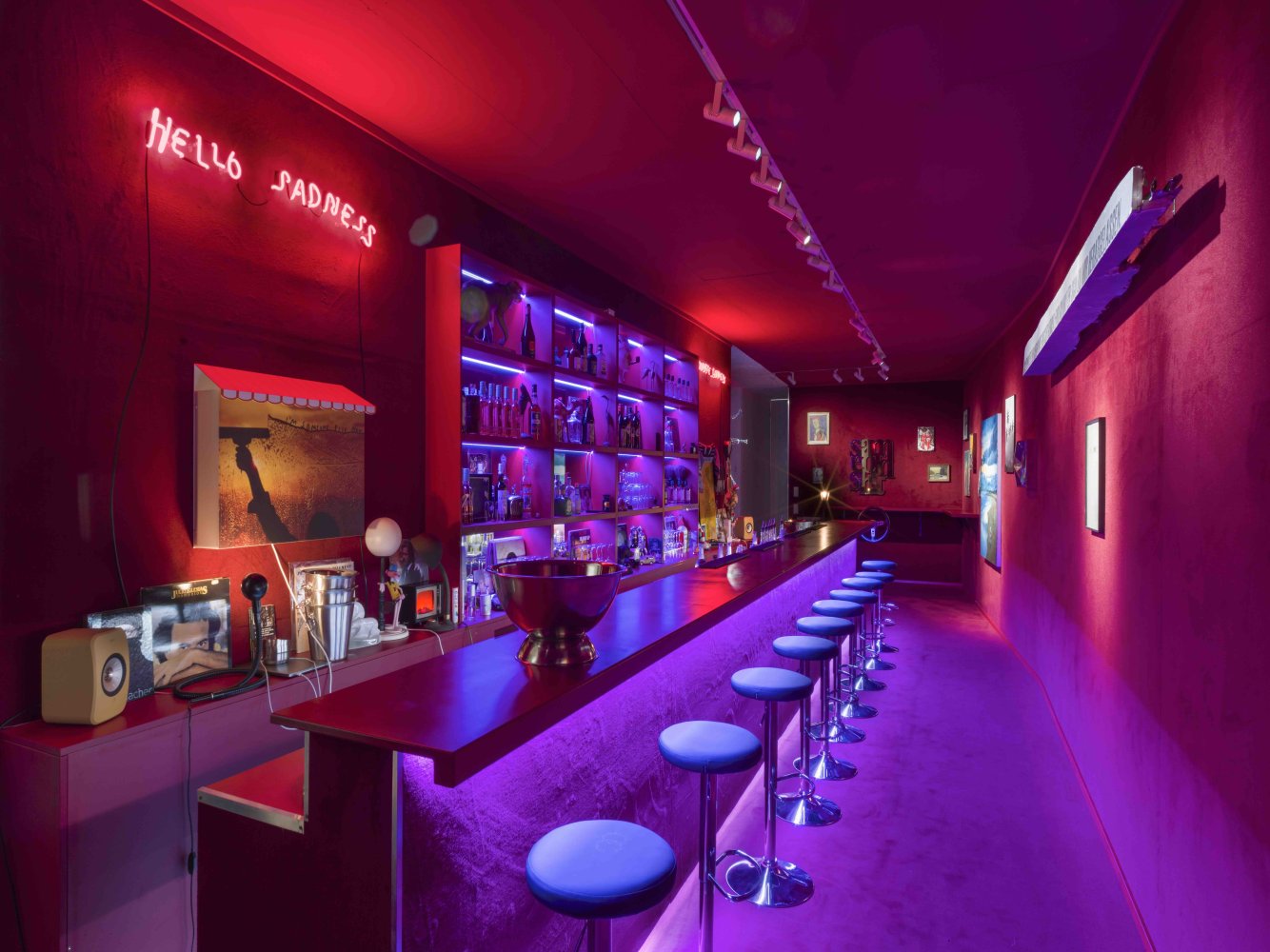 Installationsansicht: langer violett-pinker Barraum mit Tresen, Hockern und Neonbeleuchtung.