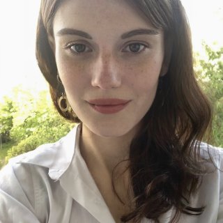 Porträtfoto einer Frau in weißer Bluse, neutraler Blick, draußen vor grünem Hintergrund.